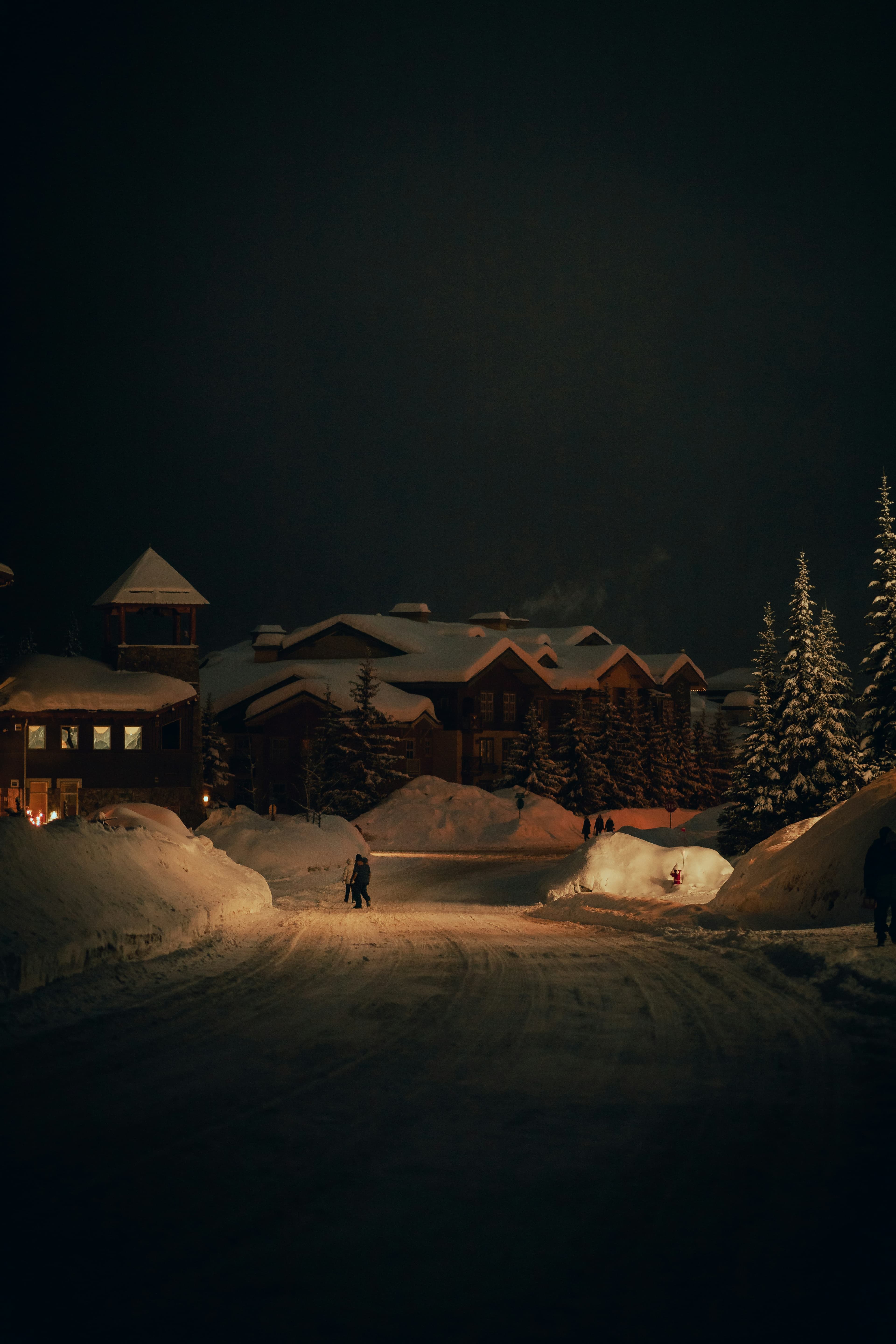 A snowy night and a street, a few houses and a person are lit up by a street light