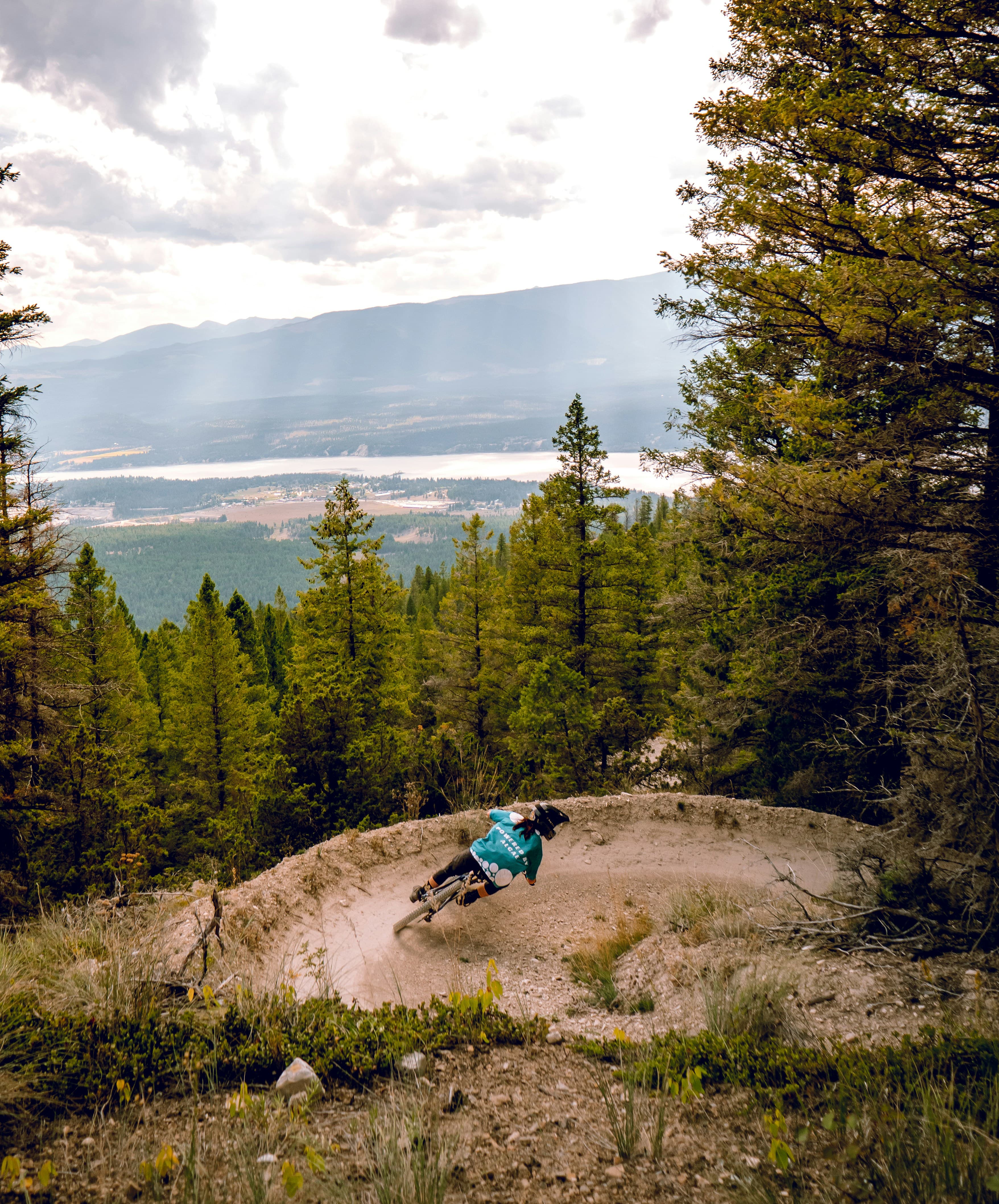 A mountain biker on a dirt berm with a view of the city, river, and surrounding mountains coming through the trees.