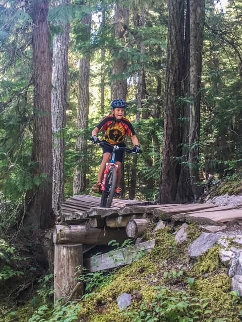 A young boy mountain biking in Whistler in the forest on a wooden ramp above the ground